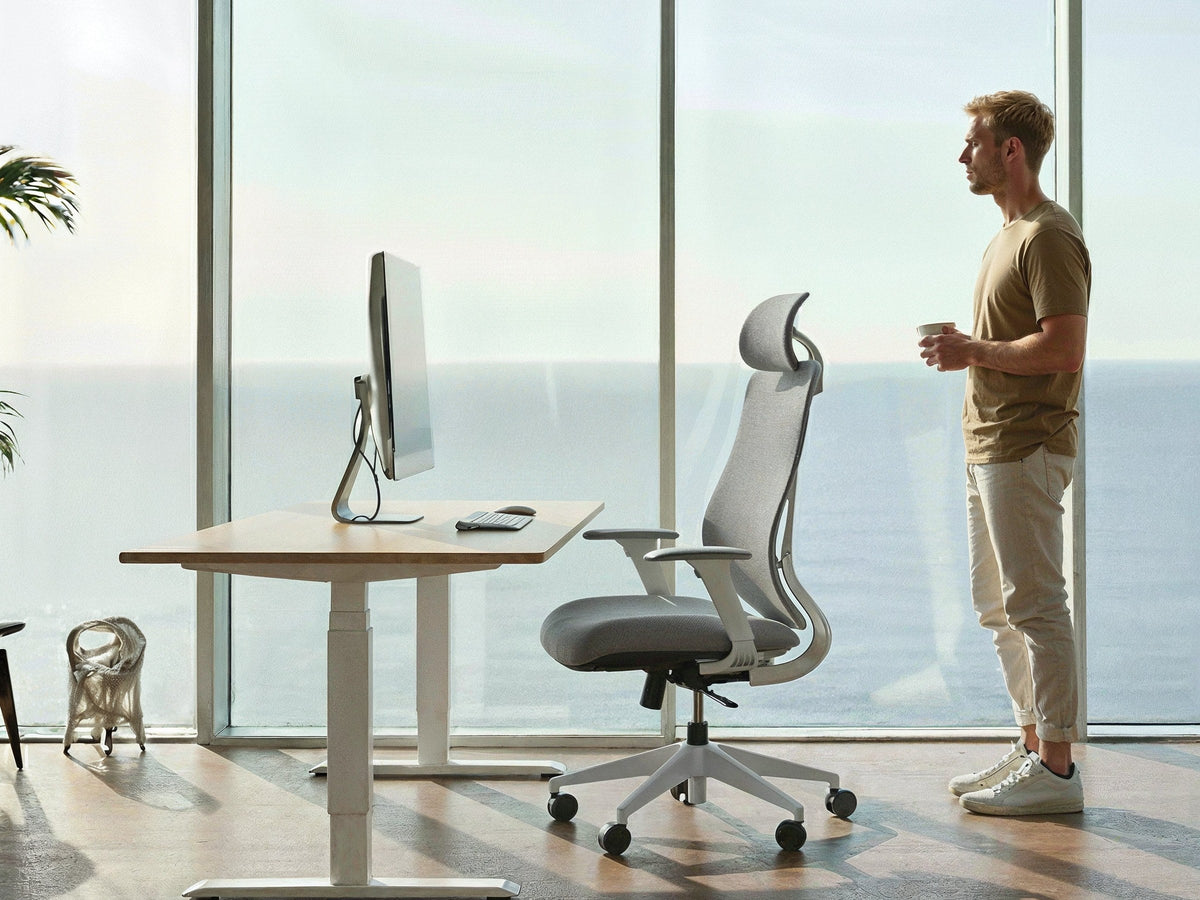 Ergonix reguliuojamo aukščio stalas ir OmniSit Pro kėdė prie didelio lango, šalia stovi vyras su puodeliu